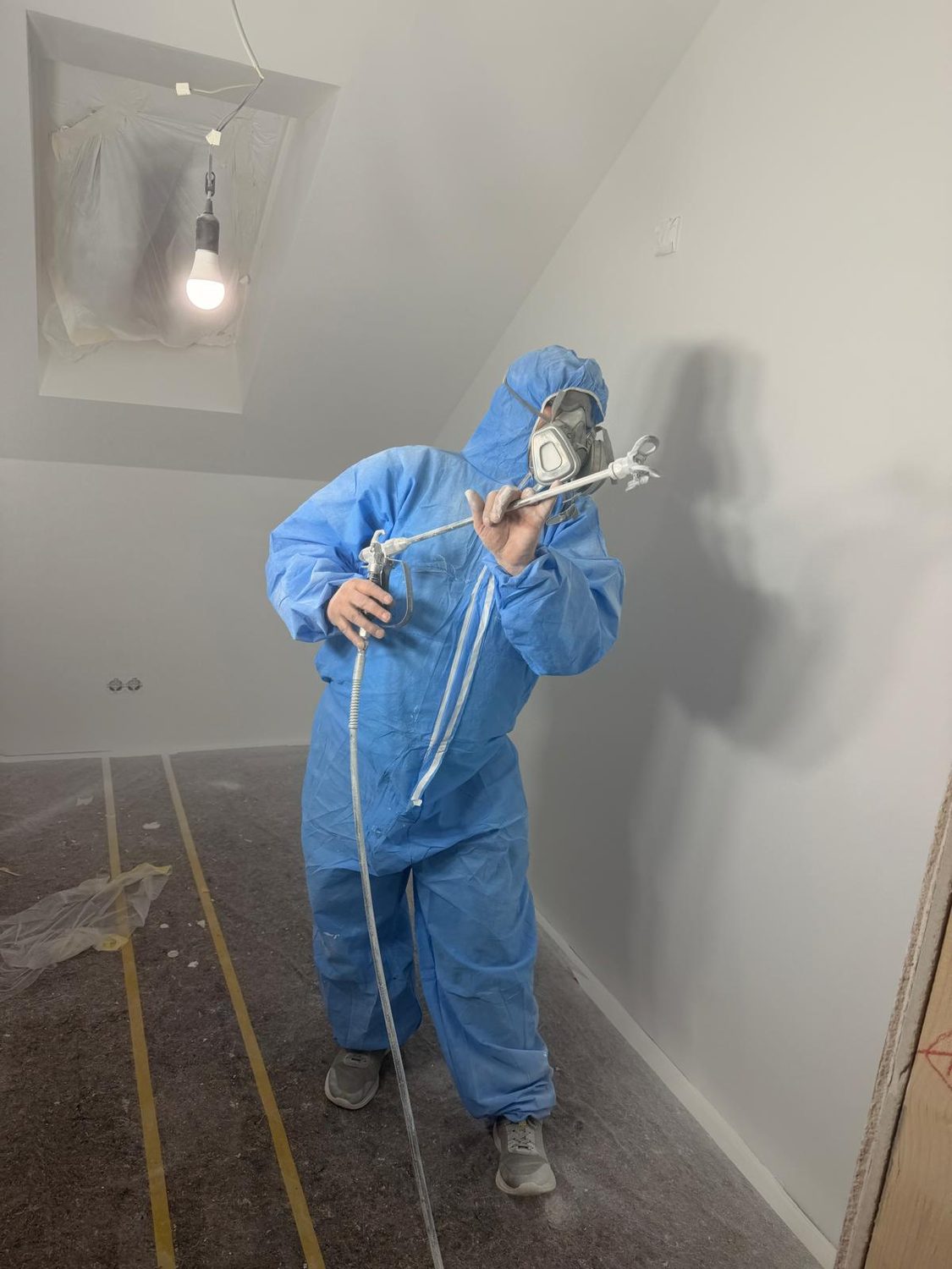 Maler von Mozart Bau trägt im Schutzanzug mit Airless-Gerät Farbe auf eine Dachschräge auf